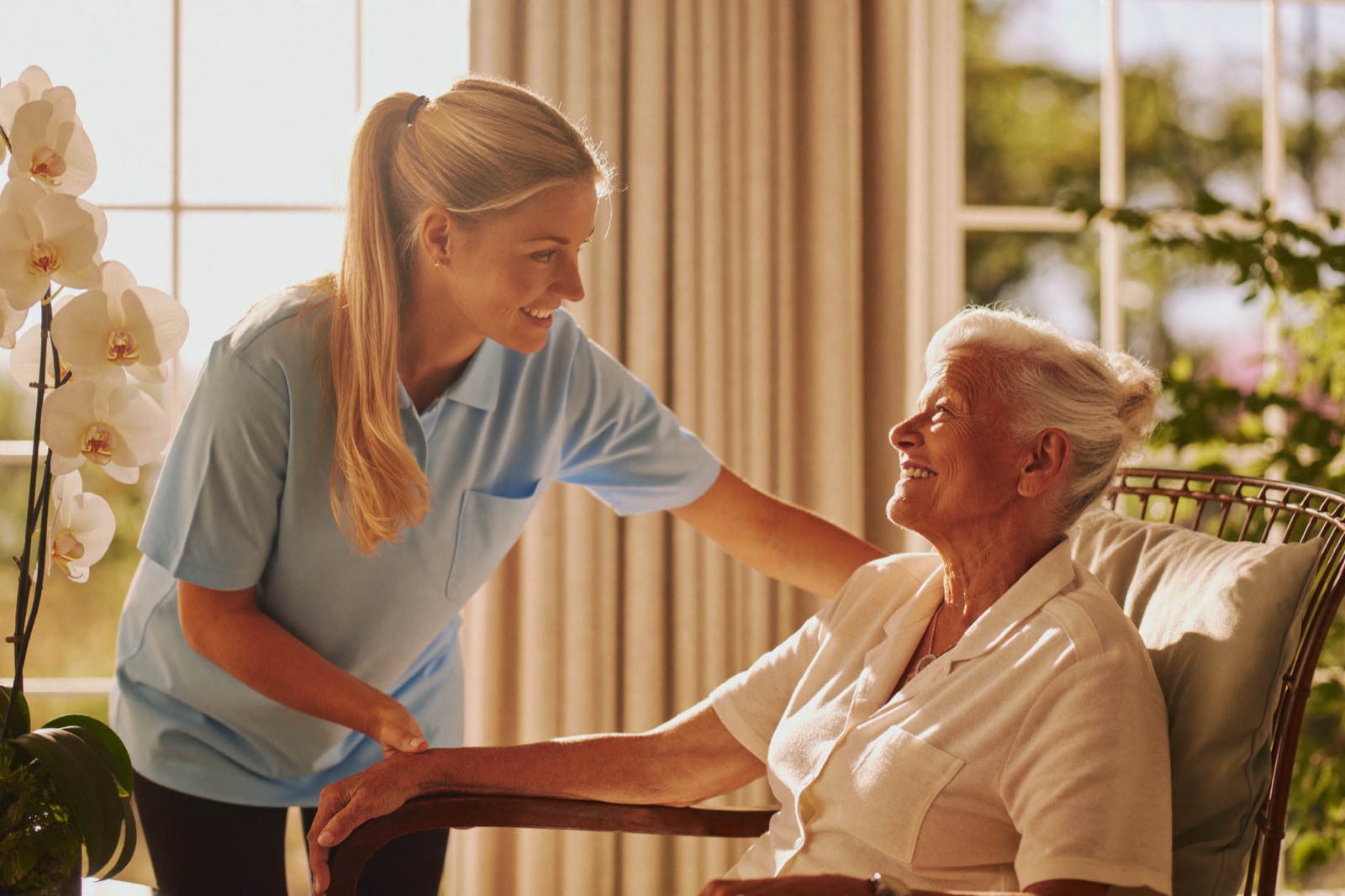 A home health aide helping a senior woman tend her orchids by a bright window