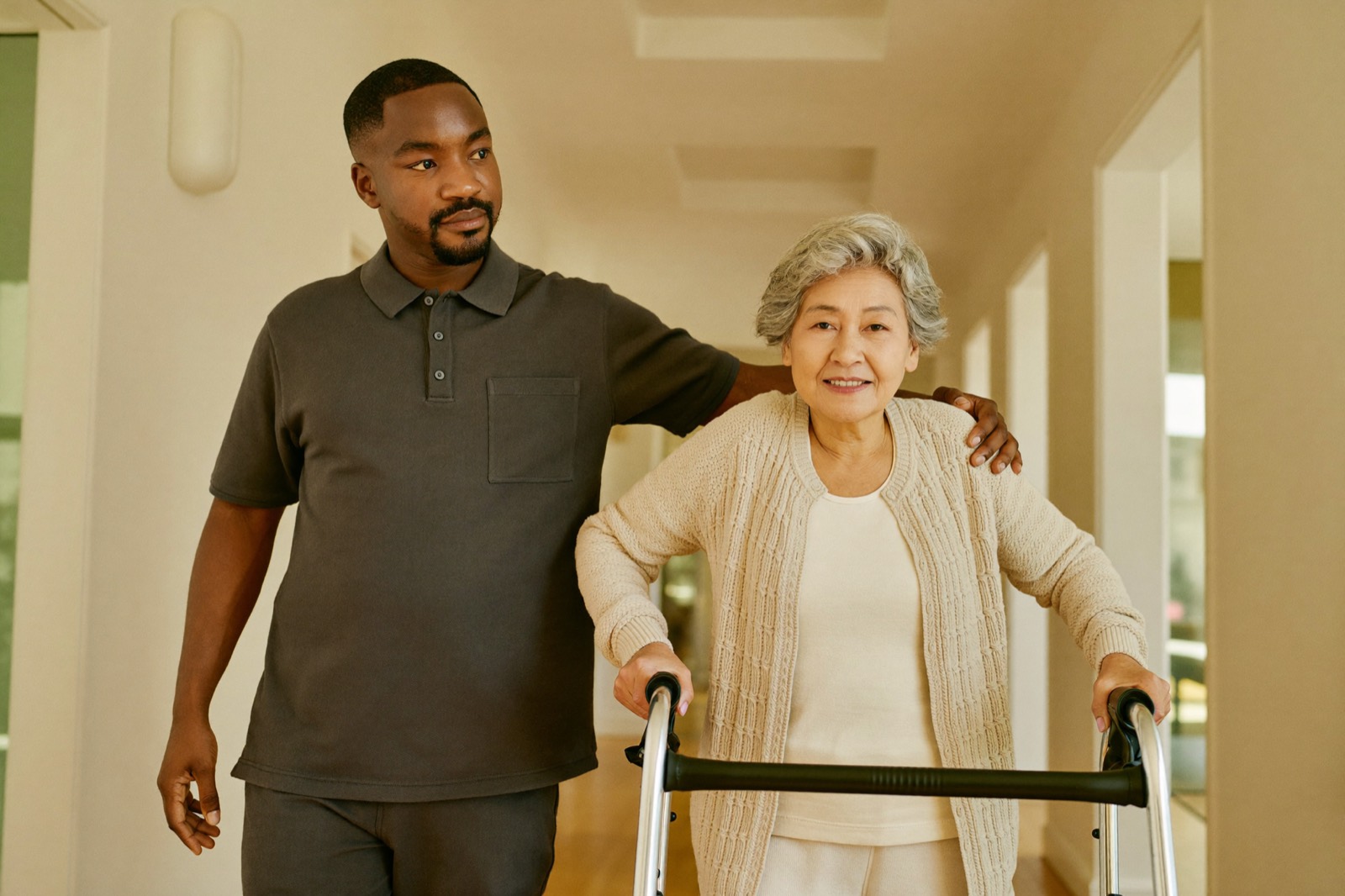 A physical therapist steadying a senior woman as she walks with a rollator in a bright hallway