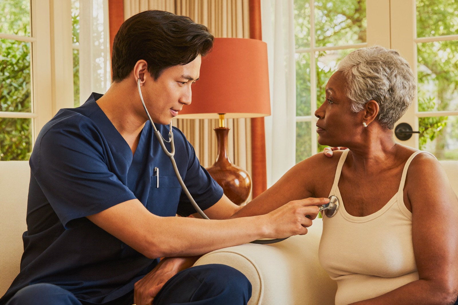 A home health nurse listening to a senior man's heart with a stethoscope in his living room
